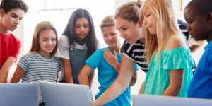 A teacher pointing to a laptop screen surrounded by her young students.